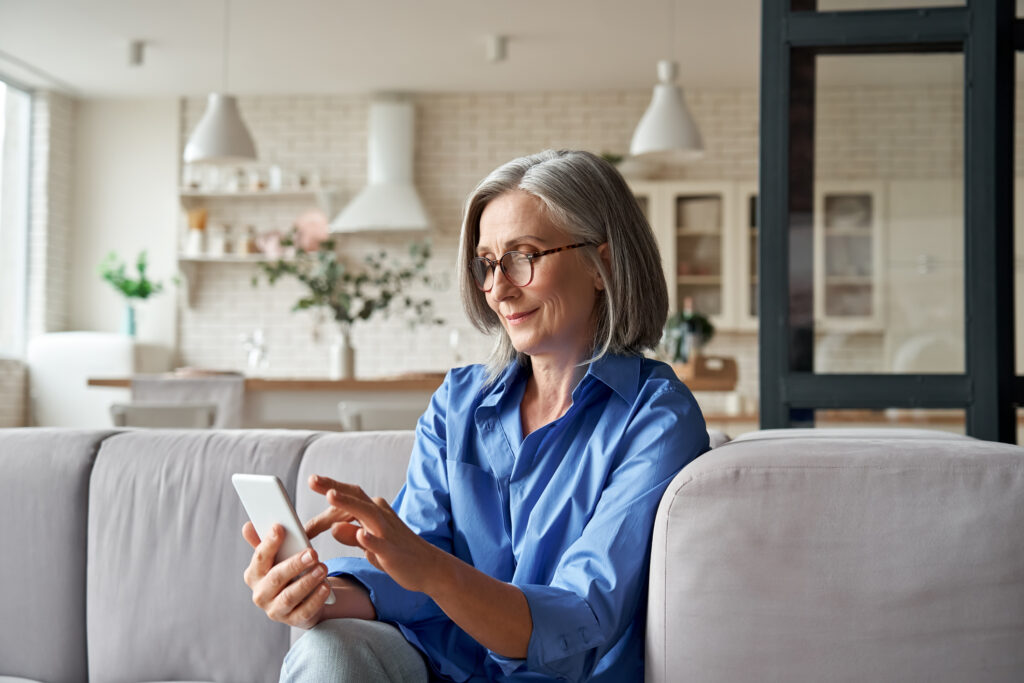 Relaxed mature old 60s woman, older middle aged female customer holding smartphone using mobile app, texting message, search ecommerce offers on cell phone technology device sitting on couch at home.